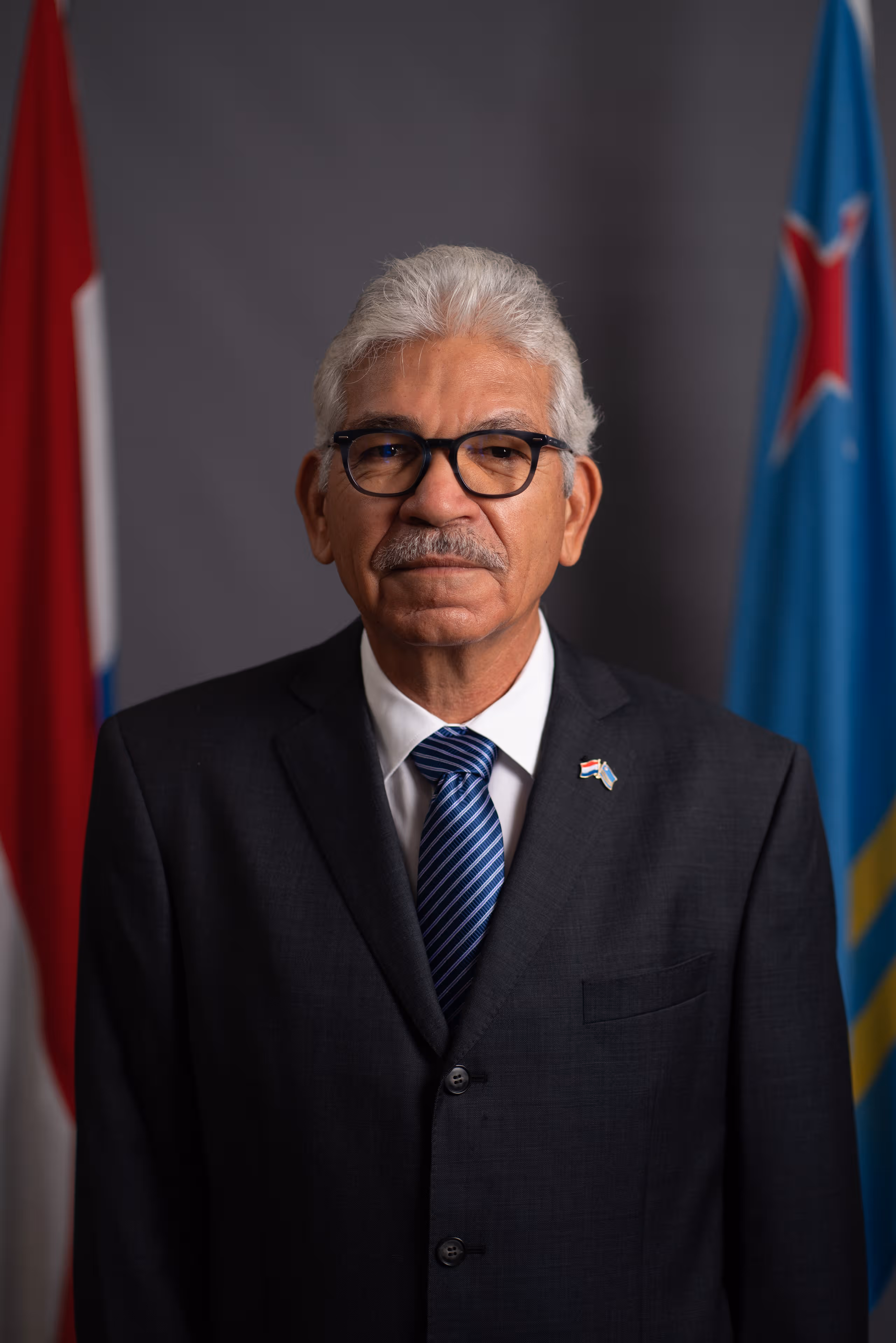 Portretfoto van Waarnemend Gouverneur Vrolijk. Grijze achtergrond met links de Nederlandse en rechts de Arubaanse vlag.