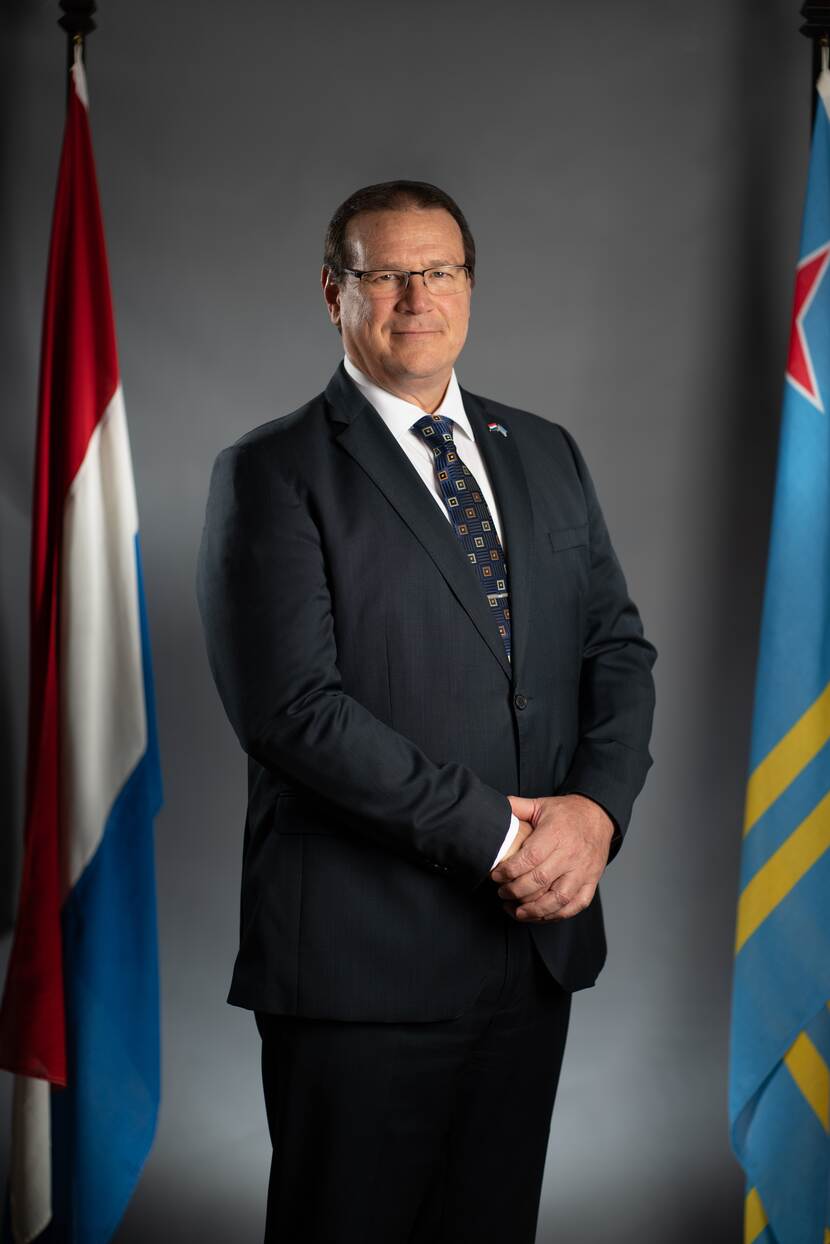 Portretfoto van Gouverneur Boekhoudt. Grijze achtergrond met links de Nederlandse en rechts de Arubaanse vlag.