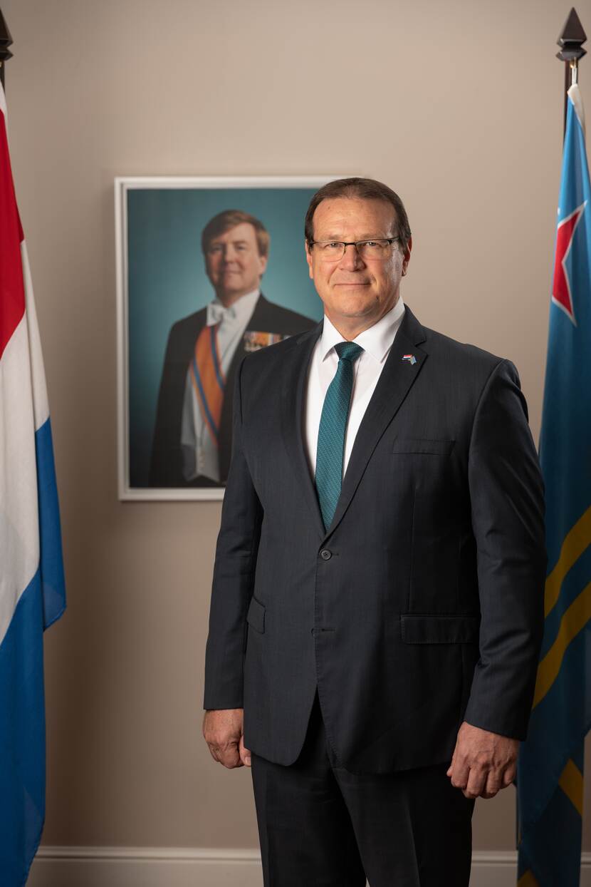 Portretfoto van Gouverneur Boekhoudt. Op de achtergrond een staatsieportret van Koning Willem-Alexander. Links van de Gouverneur staat de Nederlandse en rechts de Arubaanse vlag.